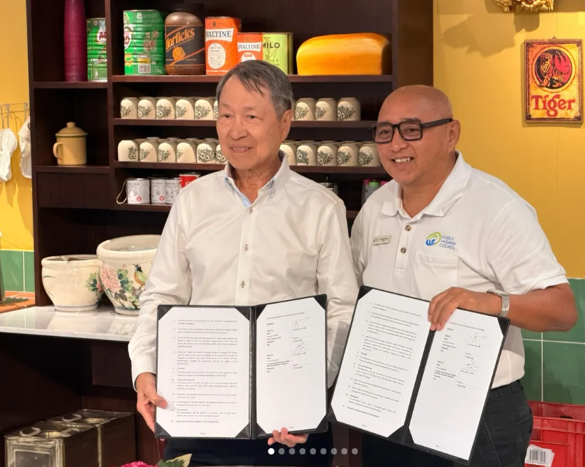 Two individuals holding up signed documents in a retro-styled setting, with one person wearing a Public Hygiene Council polo shirt, marking a formal agreement or partnership signing.